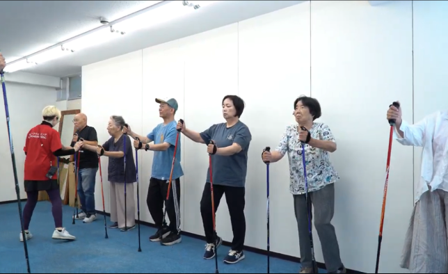 教室で参加者が横一列に並び、専用ポールを持って運動する様子